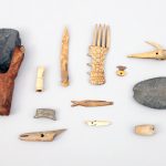 A selection of artifacts and tools excavated from the Kuukpak archaeological site in the 1980s. Artifacts include stone adze, decorated comb, fishing lures, harpoon heads, stone net sink, needle case, awl, bird bunt, and copper ulu pendant.