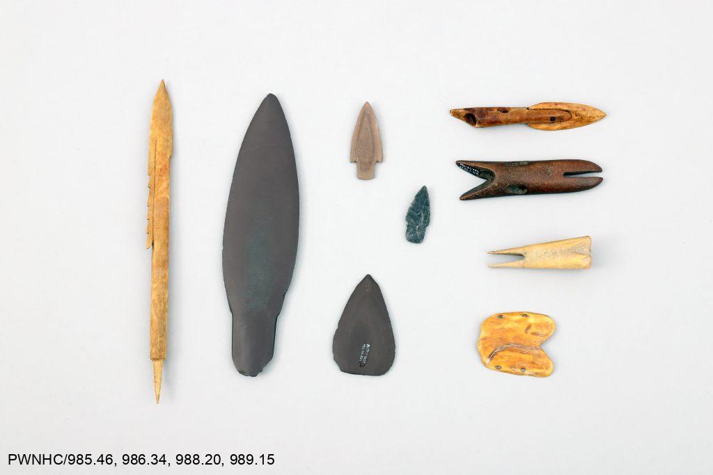Bone arrowhead, slate lanceolate blade, stone projectile points and harpoon blades, harpoon heads, bird bunt, and a wrist guard.