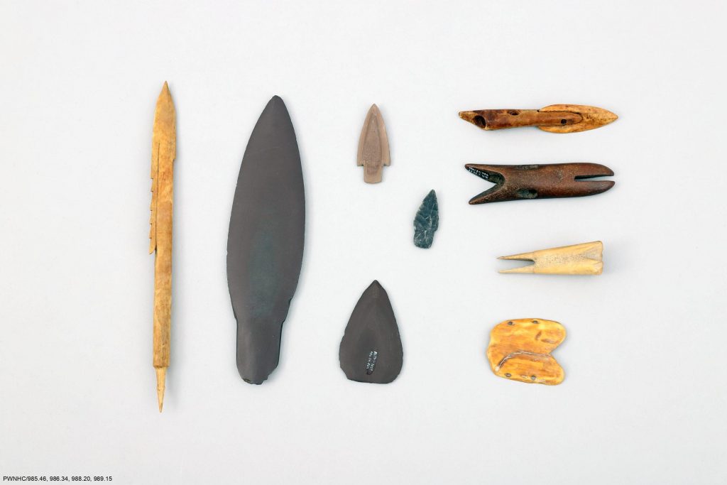 Bone arrowhead, slate lanceolate blade, stone projectile points and harpoon blades, harpoon heads, bird bunt, and a wrist guard.