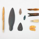 Bone arrowhead, slate lanceolate blade, stone projectile points and harpoon blades, harpoon heads, bird bunt, and a wrist guard.