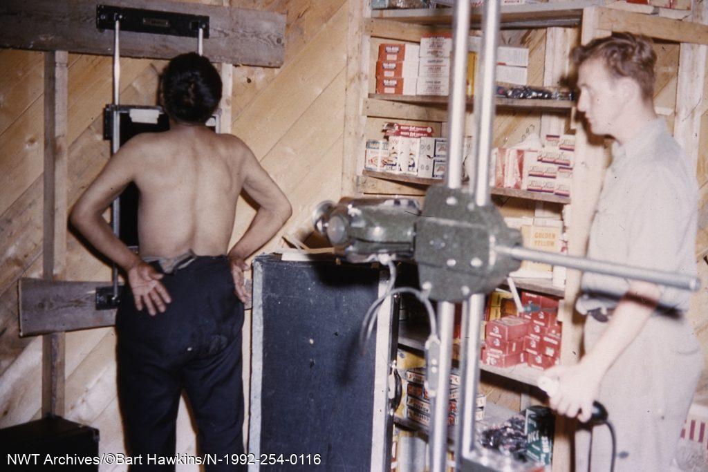 Un technicien d’Ottawa réalise la radiographie d’un homme dans le magasin de la Compagnie de la Baie d’Hudson, à Lutselk’e, en 1955. De nombreuses personnes des TNO qui souffraient de la tuberculose étaient envoyées à Edmonton, habituellement l’hôpital Charles  Camsell, où elles restaient pendant plusieurs années pour se rétablir. 